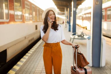 Tren istasyonunda tren vagonuna binmek için beklerken cep telefonuyla konuşan mutlu Avrupalı bayan gezgin.