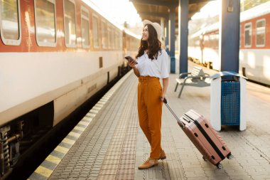 Cep telefonunu kullanan mutlu bayan vagon numarasına bakıyor, vagon sayısını kontrol ediyor ve trende bekliyor, platformda duruyor, tam uzunlukta.