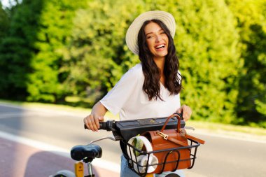 Heyecanlı bayan mutluluk saçıyor, parkta elektrikli bisikletle duruyor, çevre dostu ulaşım hizmetini paylaşıyor.