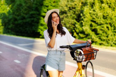 Cep telefonuyla konuşan güzel Avrupalı bir bayan, parkta elektronik bisiklet sürerken elektrikli bisikletin yanında duruyor.