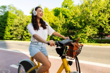 Kulaklıklı şık bir bayan şehir parkında elektrikli bisiklet sürüyor, müzikten ve yaz yürüyüşünden zevk alıyor.