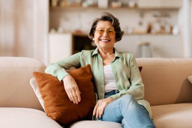 Mutlu, kendinden emin, yastığa yaslanmış, evdeki rahat kanepeye oturmuş ve kameraya gülümseyen yaşlı kadın portresi. Mutlu emeklilik kavramı