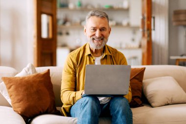 Dizüstü bilgisayar kullanan, internette gezinen, gülümseyen, evdeki kanepede otururken online çalışan mutlu kıdemli bir adam. Yaşlılıkta serbest çalışma