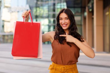 Kese kağıdını gösteren, kameraya gülümseyen, açık havada duran, maket yapan heyecanlı genç bayan. Tüketim ve ticaret kavramı