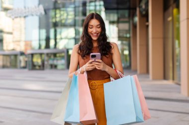 İnternetten alışveriş. Akıllı telefonu ve parlak kağıt torbaları olan mutlu bir bayan mobil uygulama ile alışveriş yapmayı seviyor, alışveriş merkezinin dışında bekliyor.