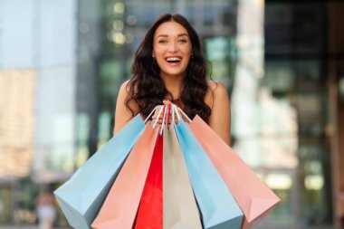 Bir sürü renkli alışveriş çantasıyla tatmin olmuş Avrupalı kadın dışarıda poz veriyor, alışverişi seven kadın kameraya gülümsüyor.