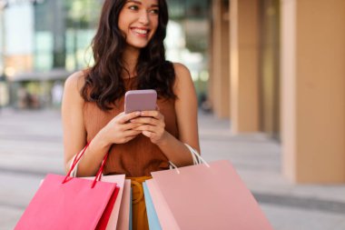 Alışveriş torbalı şık bir bayan cep telefonu kullanıyor, yakın plan. Alışveriş merkezinde alışveriş yapan bir kadın, dışarıda dikilen, yan bakan ve gülümseyen, boş alan