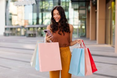 Mutlu bayan online alışveriş için akıllı telefon kullanıyor ya da taksi çağırıyor, parlak alışveriş torbaları tutuyor, mevsimlik satışların keyfini çıkarıyor.