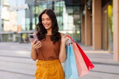 Mutlu genç bir kadın alışveriş merkezinin yakınında dururken cep telefonu kullanıyor ve elinde kese kağıtlarıyla alışverişin tadını çıkarıyor.