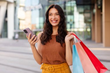 Akıllı telefonunu kullanan mutlu Avrupalı bir kadın dışarıda alışveriş torbalarıyla yürürken, taksi çağırırken, başka tarafa bakarken ve gülümserken.
