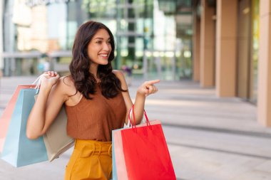 Bir sürü renkli alışveriş torbası olan alışverişçi kadın dükkanın camını işaret ediyor, alışveriş merkezinin önünde yürüyor. Şok edici bir teklif, reklam ve alışveriş