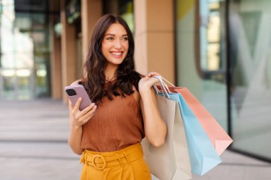 Heyecanlı müşteri bayanın elinde akıllı telefon ve renkli alışveriş çantaları var. Alışveriş merkezinin yakınında duruyorlar. Alışveriş ve e-ticaret.