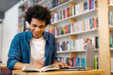 Üniversite kütüphanesinde kitap okuyan siyah bir üniversite öğrencisi dizüstü bilgisayarla oturmuş, proje bilgilerini arıyor, uzayı kopyalıyor.