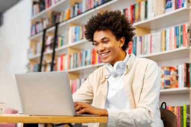 Happy Black erkek üniversite öğrencisi konferansa hazırlanıyor, kolej kütüphanesinde okurken dizüstü bilgisayar kullanıyor, modern kablosuz teknoloji ve internet kaynakları kullanıyor.