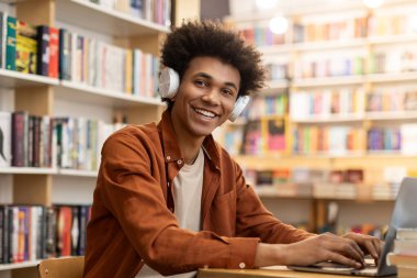 Kolej kütüphanesinde dizüstü bilgisayar kullanan, masada oturup kameraya gülümseyen, kulaklıklı Afrikalı Amerikalı bir öğrencinin portresi.