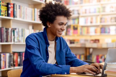 Üniversite kütüphanesinde dizüstü bilgisayarla çalışan mutlu Afrikalı Amerikalı öğrenci, kitaplıkların arasında masa başında oturuyor, uzaktan kumandalı konferans veriyor.