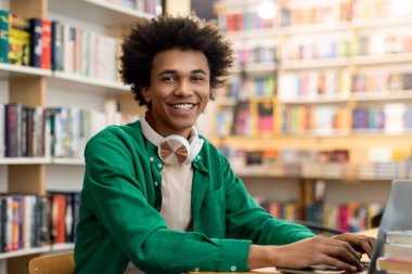 Mutlu Afrikalı Amerikalı erkek öğrenci kütüphanede oturmuş kameraya gülümsüyor, online ders alıyor ya da derse hazırlanıyor.