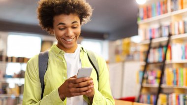 Akıllı telefonunu kullanan, sosyal ağlarda arkadaşlarıyla sohbet eden ve kütüphanede durup dostça gülümseyen mutlu Afrikalı Amerikalı öğrenci.