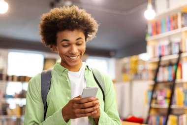 Akıllı telefonunu kullanan, sosyal ağlarda arkadaşlarıyla sohbet eden ve kütüphanede durup dostça gülümseyen mutlu Afrikalı Amerikalı öğrenci.