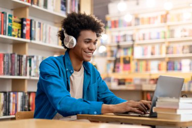 Üniversite kütüphanesinde dizüstü bilgisayarla çalışan mutlu Afrikalı Amerikalı öğrenci, kitaplıkların arasında masa başında oturuyor, uzaktan kumandalı konferans veriyor.
