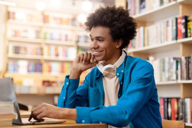 Mutlu Latin erkek öğrenci kampüs kütüphanesinde okuyor, dizüstü bilgisayar kullanıyor, derse hazırlanıyor ya da proje için bilgi topluyor