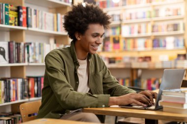 Üniversite kütüphanesinde dizüstü bilgisayarla çalışan mutlu Afrikalı Amerikalı öğrenci, kitaplıkların arasında masa başında oturuyor, uzaktan kumandalı konferans veriyor.