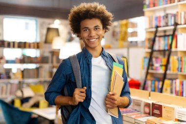 Sırt çantası takan ve üniversite kütüphanesinde kameraya gülümserken elinde bir yığın kitap tutan mutlu Afrikalı Amerikalı adam. Üniversitede yeni akademik dönem