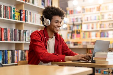 Üniversite kütüphanesinde dizüstü bilgisayarla çalışan mutlu Afrikalı Amerikalı öğrenci, kitaplıkların arasında masa başında oturuyor, uzaktan kumandalı konferans veriyor.