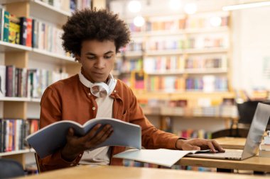 Üniversite kütüphanesinde oturup dizüstü bilgisayar kullanan, ders ya da sınava hazırlanan siyah öğrenci kitap okuyor.
