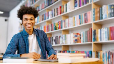 Kablosuz kulaklık takan, kütüphanede oturmuş notlar alırken kameraya gülümseyen heyecanlı Latin öğrenci portresi.