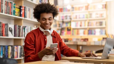 Üniversitenin kütüphanesinde masasında otururken cep telefonu kullanan pozitif siyah erkek öğrenci laptopta sınavlara hazırlanırken mesaj atıyor.