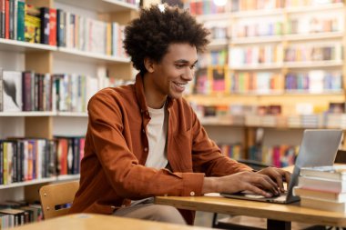 Üniversite kütüphanesinde dizüstü bilgisayarla çalışan mutlu Afrikalı Amerikalı öğrenci, kitaplıkların arasında masa başında oturuyor, uzaktan kumandalı konferans veriyor.