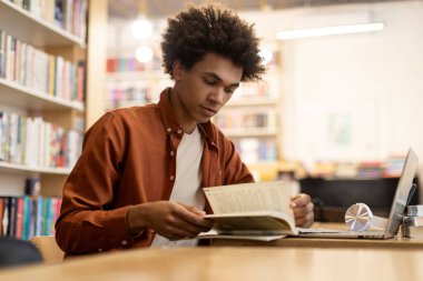 Etrafı kitaplarla çevrili çalışkan bir Afrikalı Amerikalı, üniversite kütüphanesi ortamında eğitici okumaya odaklanmış, dizüstü bilgisayarla masada oturuyor.
