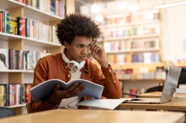 Kampüs kütüphanesindeki masada oturan düşünceli siyah öğrenci, proje ya da ödev yapıyor, defterini tutuyor ve laptop ekranına bakıyor.