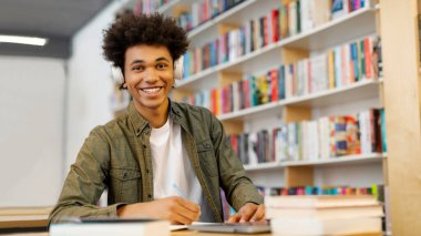 Kablosuz kulaklık takan, kütüphanede oturmuş notlar alırken kameraya gülümseyen heyecanlı Latin öğrenci portresi.