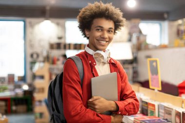 Sırt çantası takan ve dizüstü bilgisayarı olan heyecanlı Afrikalı Amerikalı çocuk, yeni akademik dönemin başında üniversite kütüphanesinde duruyor.