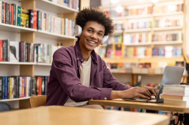 Kolej kütüphanesinde dizüstü bilgisayar kullanan, masada oturup kameraya gülümseyen, kulaklıklı Afrikalı Amerikalı bir öğrencinin portresi.