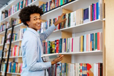 Kütüphaneden kitap alan mutlu Latin öğrenci öğrenimi için bilgi ve akademik kaynaklar arıyor, kameraya bakıyor ve gülümsüyor.