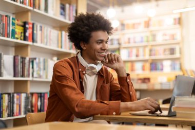 Mutlu Latin erkek öğrenci kampüs kütüphanesinde okuyor, dizüstü bilgisayar kullanıyor, derse hazırlanıyor ya da proje için bilgi topluyor