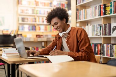 Üniversite kütüphanesinde dizüstü bilgisayar kullanan ve teste hazırlanan ya da Webinar izleyen mutlu Afrikalı Amerikalı öğrenci.