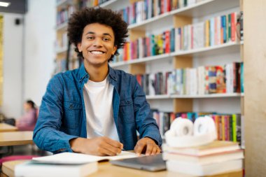 Kütüphanedeki mutlu Latin erkek öğrencinin portresi, eğitime dalmış kitapların arasında oturuyor ve kameraya gülümsüyor, boşluğu kopyalıyor.