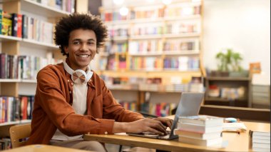 Mutlu Afrikalı Amerikalı erkek öğrenci kütüphanede oturmuş kameraya gülümsüyor, online ders alıyor ya da derse hazırlanıyor.