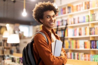 Sırt çantası takan ve elinde kitaplarla üniversite kütüphanesinde kameraya gülümseyen mutlu Afrikalı Amerikalı adamın portresi.