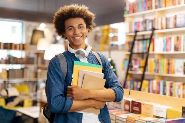 Sırt çantalı mutlu Latin öğrenci portresi. Elinde kitaplarla üniversite kütüphanesinde durup kameraya gülümsüyor.