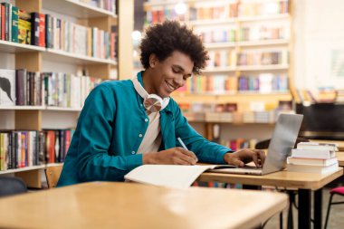 Üniversite kütüphanesinde dizüstü bilgisayar kullanan ve teste hazırlanan ya da Webinar izleyen mutlu Afrikalı Amerikalı öğrenci.