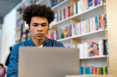 Kütüphanede dizüstü bilgisayar kullanan, kablosuz kulaklık takan ve online konferans ya da webinar dinleyen konsantre siyahi adam. Teknoloji, eğitim kavramı