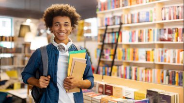 Sırt çantalı, sırtüstü gülümseyen, kitaplarını kütüphanede tutarken başarı saçan, kitapların önünde duran mutlu siyahi öğrenci.