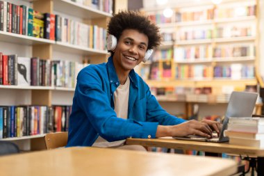 Kolej kütüphanesinde dizüstü bilgisayar kullanan, masada oturup kameraya gülümseyen, kulaklıklı Afrikalı Amerikalı bir öğrencinin portresi.