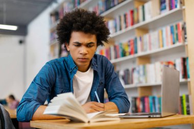 Kitap bilginin kaynağıdır. Kütüphane masasında kitap okuyan, yeni malzemeler okuyan düşünceli siyah bir çocuk.