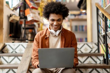 Üniversite kütüphanesinde merdivenlerde oturup ders arası verip ders için bilgi arayan mutlu siyahi öğrenci.